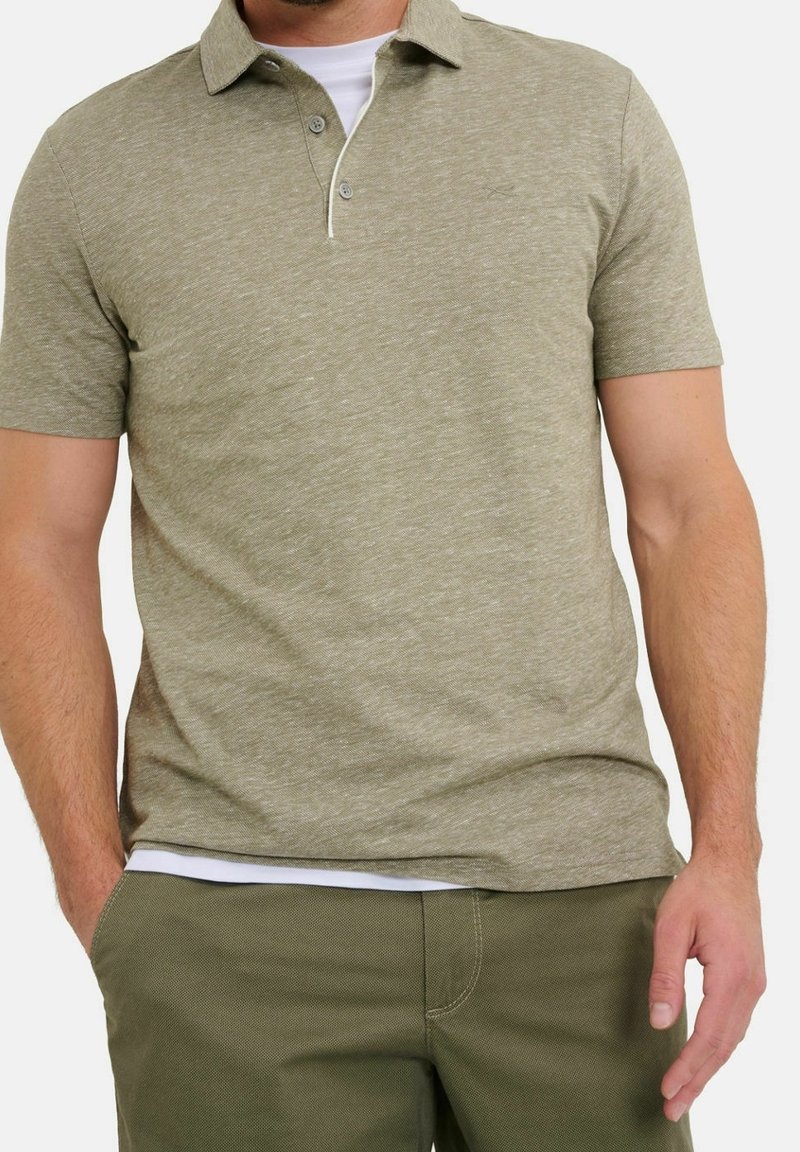 Homme portant un polo manches courtes olive clair sur un t-shirt blanc avec un pantalon vert olive, une main dans la poche et l'autre détendue.