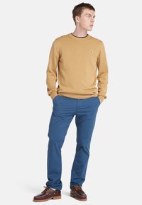 Mosterdkleurige trui met geribbelde manchetten en onderkant, gecombineerd met blauwe chino's en bruine veterschoenen. Opmerkelijk logo op de trui.
