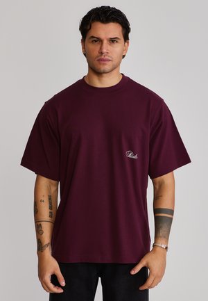 Mann mit dunklem Haar, der ein lockeres dunkelrotes T-Shirt mit einem kleinen weißen Schriftzug auf der Brust trägt und sichtbare Tattoos auf beiden Armen hat.