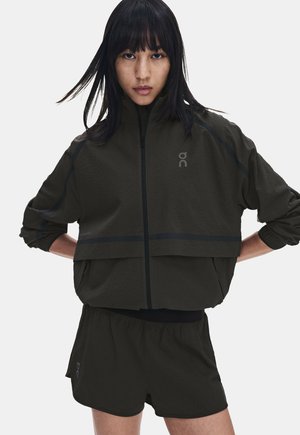 Femme aux longs cheveux foncés portant une veste noire zippée et un short assorti, debout les mains sur les hanches contre un fond uni.