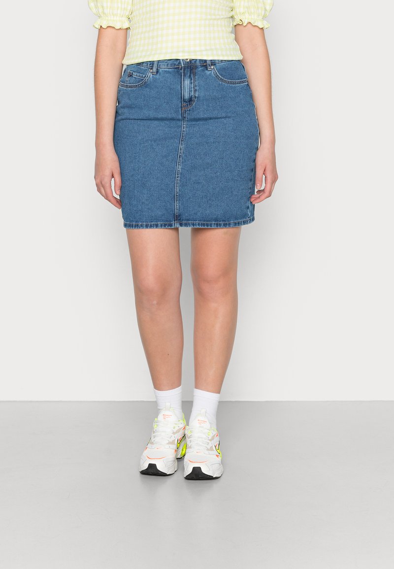 Saia de ganga em azul médio, com forma ajustada e bainha desfiada. Combinada com ténis brancos e meias. O fundo é branco liso.