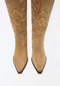Tan suede cowboy boots with pointed toes, intricate stitching patterns, and metallic studs along the side, featuring a mid-calf height design.