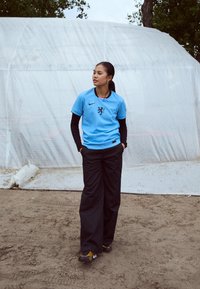 Hele halli Nike'i jersy, millel on lõvilogo, on ühtlustatud mustade laiade pükste ja mustade jalanõudega. Taustal on valge telk.