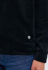 Zwarte corduroy overhemd met verticale ribbels, voorzien van lange mouwen en een ronde zoom. Een klein metallic logo is zichtbaar nabij de onderkant.