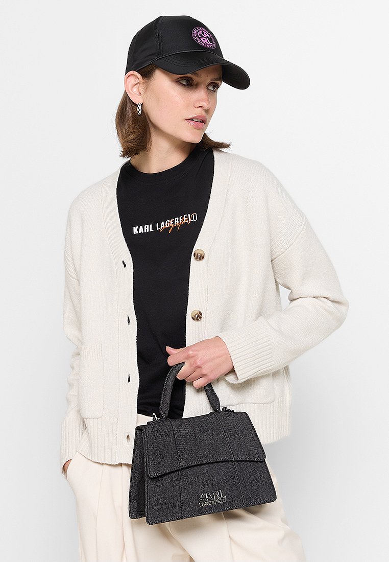 Sac à main noir texturé avec un design structuré et un rabat à l'avant, arborant un logo. Le modèle porte un cardigan crème et une casquette noire.