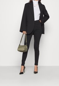 Svart oversized blazer över en figursydd vit polotröja, kombinerad med svarta skinnybyxor och högklackade skor. Håller en olivgrön handväska.