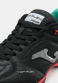 Scarpa sportiva nera da interno con texture in pelle, accenti grigi e suola rossa. Caratterizzata da una linguetta imbottita e lacci neri piatti. Dettagli di design testurizzati.
