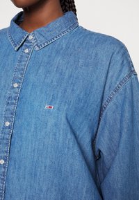 Detailní záběr na osobu oblečenou v modré denimu s knoflíky, se malým červeným, bílým a modrým logem značky na hrudi, a viditelnými zapletenými vlasy.