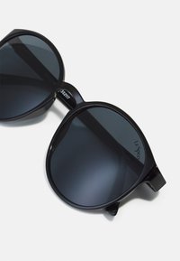 Černé oversized sluneční brýle s tmavými skly, silným rámem a hladkým povrchem. Na vnitřní části nožičky mají branding "Le Specs".