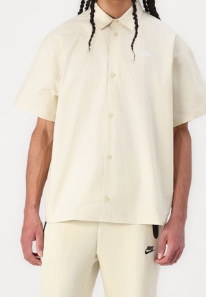 Person trägt eine cremefarbene kurzärmelige Hemdjacke und passende Hosen mit schwarzen Nike-Logos auf Hemd und Hosen.