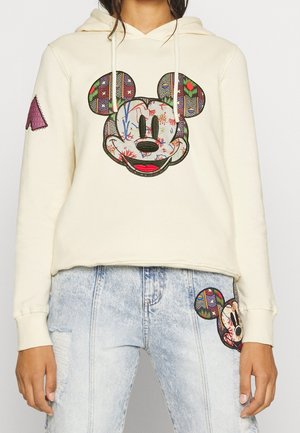 Personne portant un sweat à capuche crème et un jean bleu clair, tous deux avec des patchs colorés en forme de tête de souris de dessin animé.