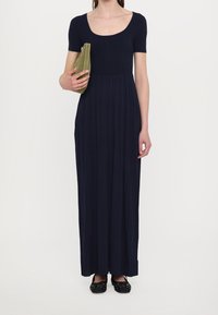 Marineblaues, geripptes Maxikleid mit kurzen Ärmeln, ausgestattet mit einem taillierten Oberteil und einem fließenden, plissierten Rock. Das Model hält eine grüne Clutch und trägt schwarze Ballerinas.