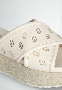 Sandali scivolati beige con cinturini incrociati caratterizzati da accenti metallici con il logo. La suola platform è testurizzata con una finitura glitter.