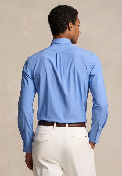 Ανοιχτό μπλε πουκάμισο με μακριά μανίκια, με δομημένο γιακά και μανσέτες με ένα κουμπί, συνδυασμένο με κρεμ παντελόνι και καφέ ζώνη.