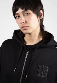 Sudadera negra con cremallera en la parte delantera y capucha ajustable con cordón. Presenta un logotipo bordado en el pecho y herrajes metálicos en la cremallera. Textura suave.