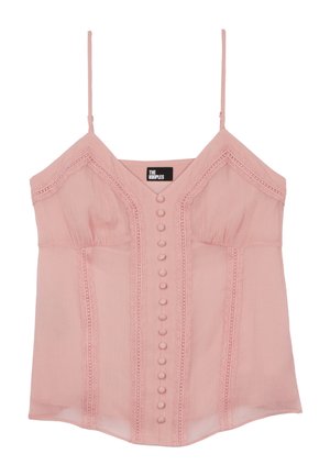 Jeune femme aux cheveux bruns lâchés portant un top rose sans manches, un pantalon blanc, des sandales compensées blanches et des bijoux en or, debout les mains dans les poches.