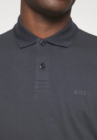 Närbild av en person som bär en mörkgrå pikétröja med ribbad krage, två knappar och en subtil "BOSS"-logotyp på bröstet.