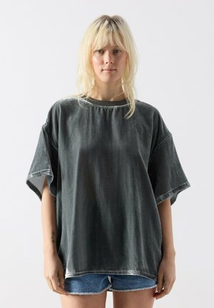 Ung person med blondt hår iført en stor, falmet sort t-shirt og blå denimshorts mod en ensfarvet hvid baggrund.