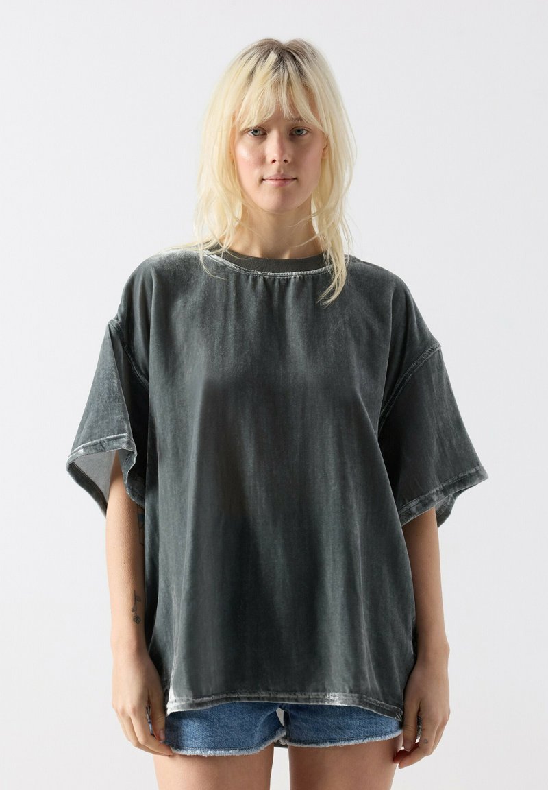 Młoda osoba o blond włosach, ubrana w za dużą, wyblakłą czarną koszulkę i niebieskie szorty dżinsowe na jednolitym białym tle.