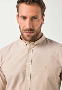 Jasnobeżowa koszula zapinana na guziki z kołnierzykiem mandaryńskim, z małym haftem na klatce piersiowej i gładkim, teksturowanym materiałem.