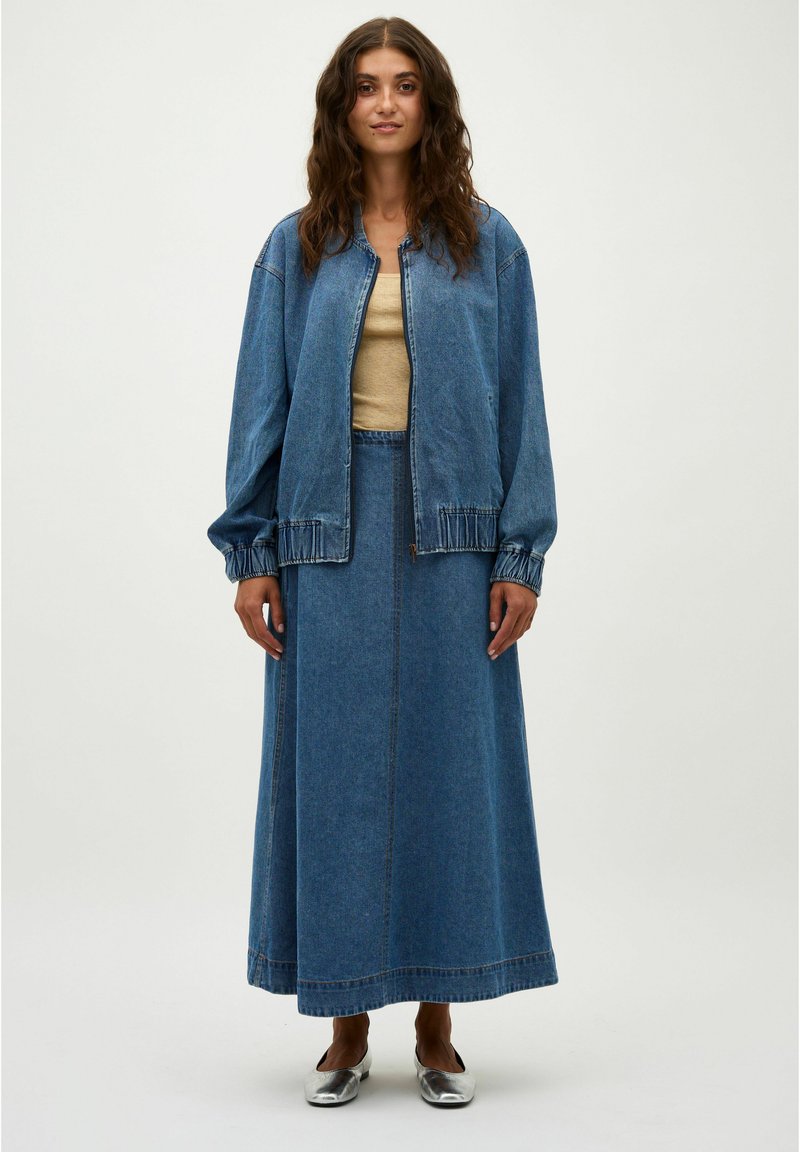 Chaqueta de mezclilla azul con puños elásticos, combinada con una larga falda de mezclilla azul. El conjunto incluye una blusa beige sencilla y zapatos planos plateados.