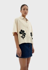 Short-sleeve, beige shirt made of fabric featuring black floral embroidery on the front, paired with a denim skirt.