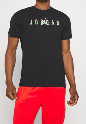Mężczyzna w czarnej koszulce Jordan z kolorowym logo i czerwonych krótkich spodenkach sportowych na jednolitym jasnym tle.