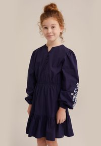 Mornarsko modra bombažna obleka s plastenim robom, napihnjenimi dolgimi rokavi in cvetličnim vezenjem na levem rokavu, ki vključuje različne odtenke modre.