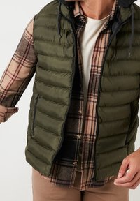 Zelená péřová vesta s horizontálním prošíváním a kapsami na zip, vrstvená přes hnědou a béžovou kostkovanou košili s světlým kulatým výstřihem.