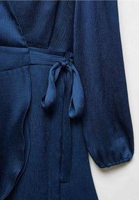 Robe portefeuille en tissu texturé bleu marine avec manches longues bouffantes et ceinture nouée à la taille sur fond blanc.