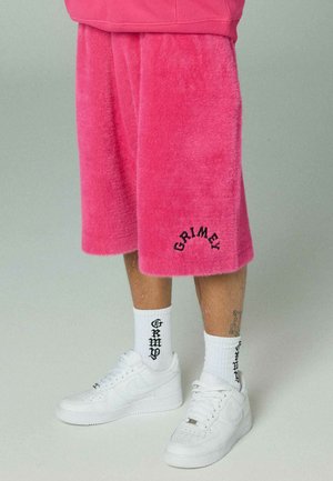 Dolne części nóg w jasnoróżowych, puszystych szortach z napisem "GRIMEY", białe skarpetki z czarnym napisem oraz białe tenisówki na neutralnym tle.