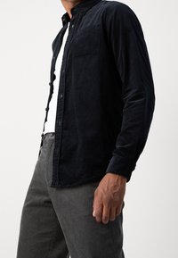 Camicia in velluto a coste blu scuro con collo button-down, taschino sul petto singolo e maniche lunghe, abbinata a pantaloni grigi a coste.