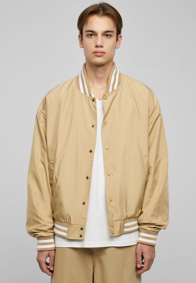 Giacca bomber tan realizzata in nylon liscio, con colletto e polsini a righe a costine, chiusura a bottoni a pressione e tasche laterali.