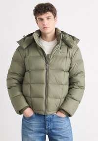 Pufferjakke i olivengrønn, med glidelås foran, avtakbar hette og quiltet design. Kombinert med blå denimjeans.
