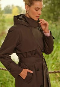 Rjav vodoodporen plašč s kapuco, velikim ovratnikom, gumbi in pasom. Ima žep na strani in gladko teksturo.