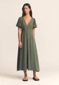 Robe maxi vert olive avec un décolleté en V profond, des manches courtes bouffantes, une fermeture à boutons sur le devant et une jupe fluide. Associée à des sandales noires simples.