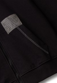 Tessuto nero con cerniera e tasca, caratterizzato da cuciture bianche e un'etichetta grigia che indica "Colore" con testo stampato in piccolo.