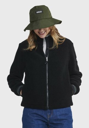 Veste en polaire noire avec fermeture éclair à l'avant, poignets côtelés et texture douce. Associée à un chapeau seau vert avec un écusson logo.