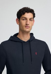 Marinblå huvtröja tillverkad av blandad bomullstextil, med snörning i huvan och en liten röd logotyp broderad på bröstet. Slät textur.