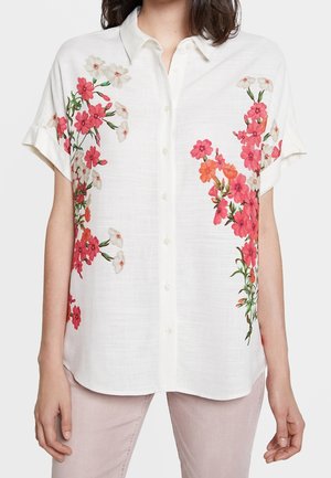 Hvid kortærmet skjorte med knapper og pink, rødt og hvidt blomsterbroderi på begge sider, båret med lyserøde bukser.