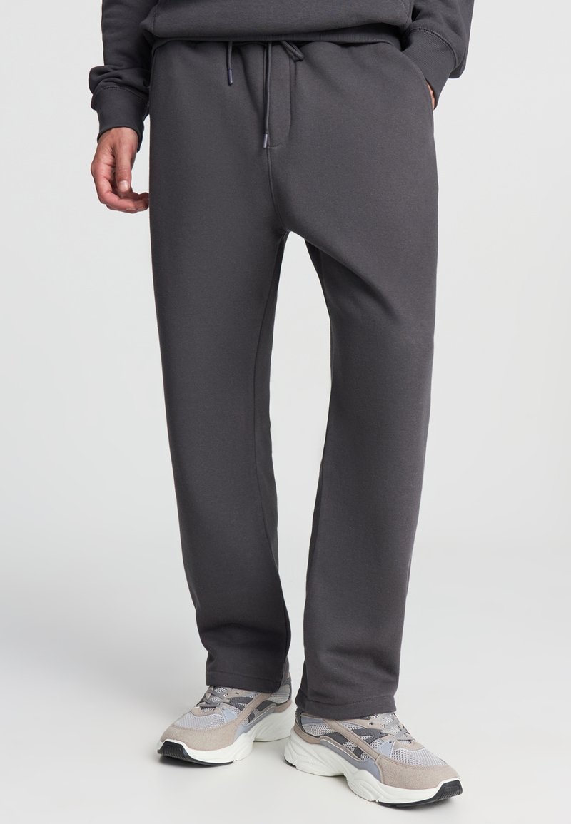 Szare dresy wykonane z miękkiego materiału, z elastycznym paskiem w talii i sznurkiem do regulacji, o luźnym kroju. Połączone z szarymi butami sportowymi.