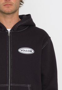 Haut à zip noir avec un logo "VOLCOM" brodé en blanc, présentant des coutures contrastantes blanches et une capuche avec cordon de serrage.