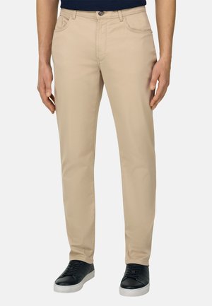 Mann trägt beige gerade geschnittene Hose und schwarze Turnschuhe, steht vor einem schlichten hellen Hintergrund.