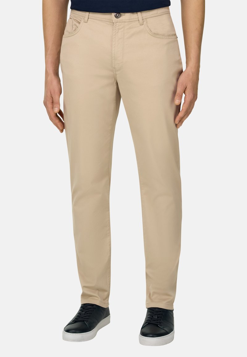 Homme portant un pantalon beige coupe droite et des baskets noires, debout devant un fond clair uni.