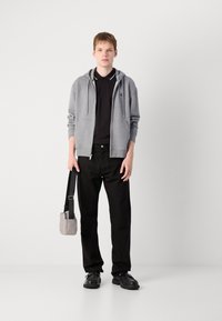 Grijze zip-up hoodie over een zwart polo, gecombineerd met zwarte broeken en zwarte schoenen. Houdt een kleine grijze crossbody tas met een schouderriem vast.