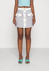 Mini-jupe en tweed aux couleurs pastel avec deux poches avant et des boutons dorés, complétée par des talons noués turquoise.