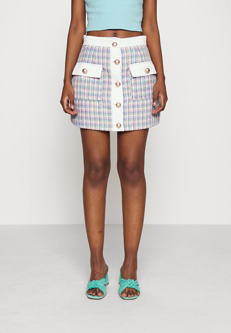 Mini-jupe en tweed aux couleurs pastel avec deux poches avant et des boutons dorés, complétée par des talons noués turquoise.