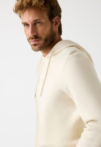 Sudadera de color crema claro con cordones ajustables, textura suave y un discreto detalle de logo en el pecho. Presenta un corte relajado y costuras cosidas.