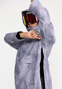 Chaqueta impermeable de color gris claro con capucha, características ajustables, bolsillo frontal y cremalleras laterales; llevada con unas coloridas gafas de esquí.