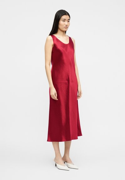 Γυναίκα με μακριά σκουρόχρωμα μαλλιά, ντυμένη με κόκκινο σατέν μίντι φόρεμα χωρίς μανίκια και λευκά μυτερά παπούτσια με χαμηλό τακούνι, που στέκεται μπροστά σε απλό φωτεινό φόντο.